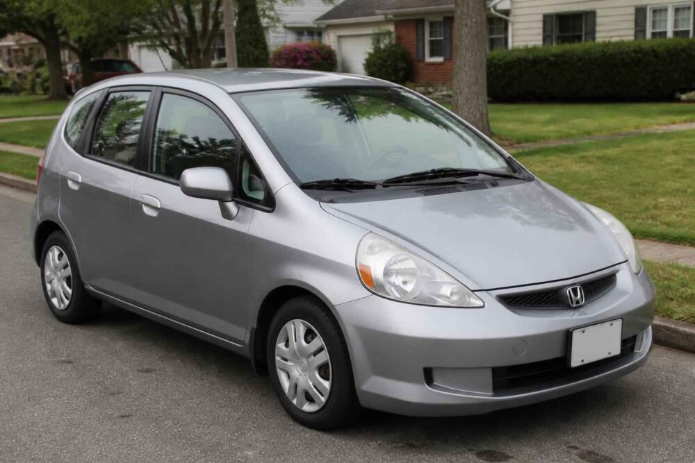 Srebrna Honda Jazz zaparkowana na osiedlowej ulicy przed domem jednorodzinnym